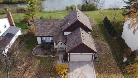 Elevated view of a single-story home with a newly installed brown architectural asphalt shingle roof on multiple steep-pitched gables. The house features brown siding and white trim and is located next to a body of water (river or lake). The foreground shows a patterned driveway and fall foliage, detailing a professional, complete roofing project in a scenic setting.